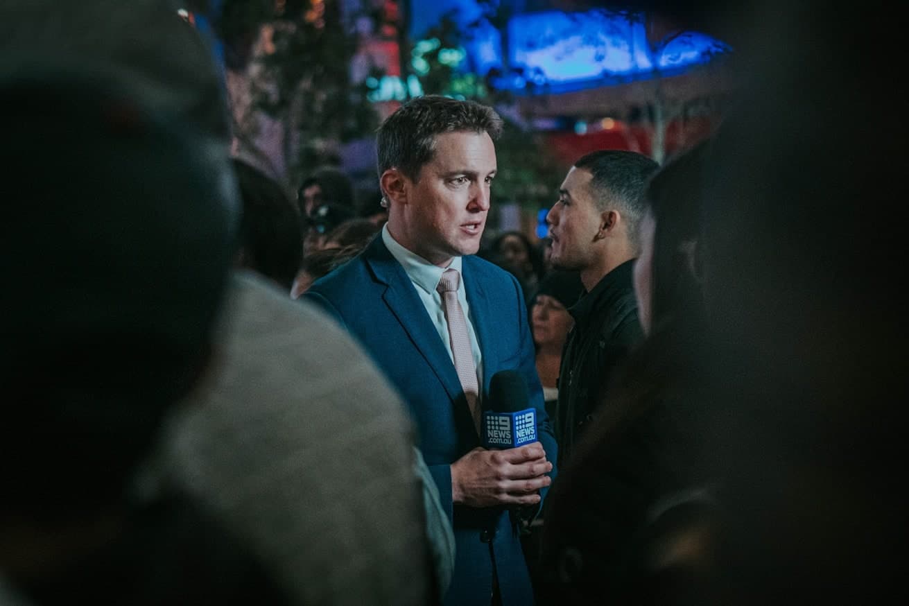 Un homme en costume, micro de télévision à la main, répond à une interview au milieu d’une foule de journalistes, lors d’une soirée.