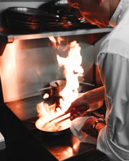 Un cuisinier en veste blanche fait flamber une préparation dans une poêle, sur un piano de cuisine professionnelle.