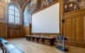 Un grand écran de projection blanc déployé devant les toiles marouflées de l’amphithéâtre, au-dessus d’une table de tribune en bois et d’un pupitre transparent.