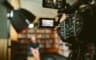 Une caméra de tournage au premier plan, son écran de contrôle cadrant une personne assise dans une bibliothèque floue.