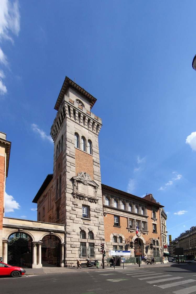 La façade de brique et de pierre de l’Institut océanographique et sa tour, à l’angle de la rue, sous un ciel bleu.